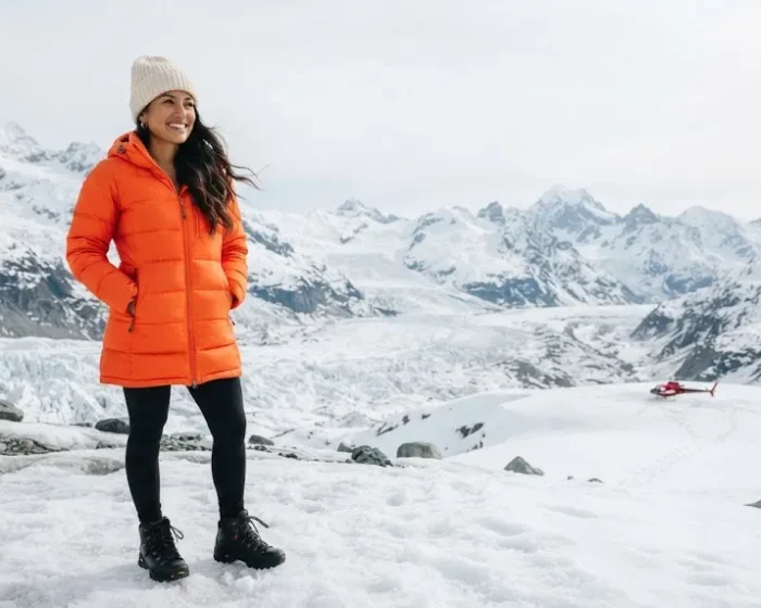 Woman in an orange puffer coat standing on a glacier in Alaska, travel capsule wardrobe outfit